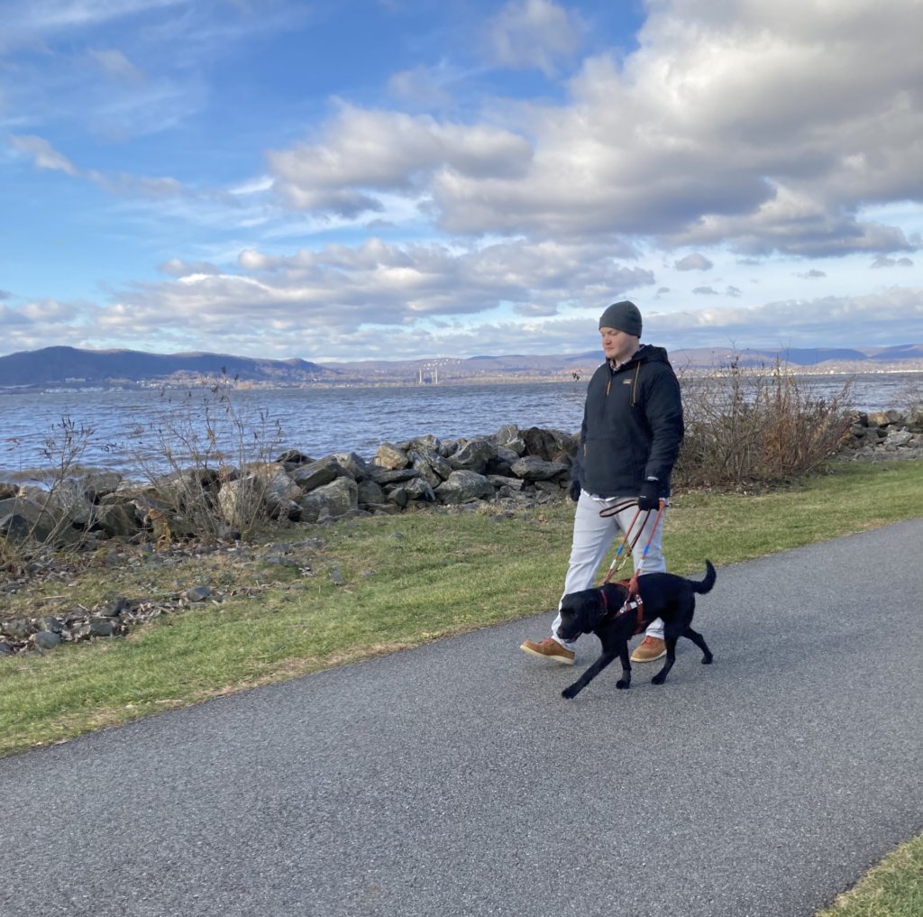 Male handler walks path along body of water with black Lab guide