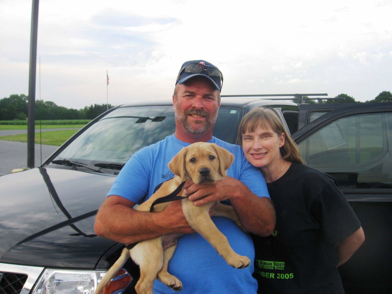Marilyn and Scott stand in a parking lot with yellow lab puppy Casper in Scott's arms.