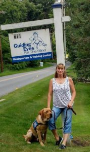 Marilyn stands while yellow lab Casper sits at her side while in his blue vest. The two pose in front of the sign by the road at the Yorktown Heights training school.