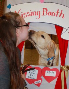 Courtney receives a kiss from yellow lab Clyde through a homemade kissing booth. On the bottom of the booth, text reads "puppy kisses for kibble with clyde" on red hearts.