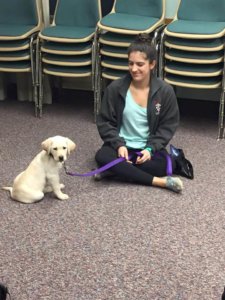 Abby sits on the ground with Fergus, a yellow lab puppy.