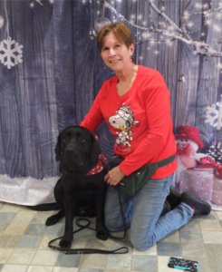 Chris kneels on the ground beside sitting black lab Orleans. The pair pose for a photo in front of a blue winter backdrop during the region holiday party.