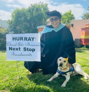 Lauren Doninger kneels in the grass while wearing a professor's graduation outfit of cap and gown with her arm on the back of laying yellow lab Meadow. Meadow is wearing her blue Guiding Eyes training Vest. The pair pose next to a sign which reads, "Hurray Liberal Arts & Sciences! Next stop, baccalaureate!"