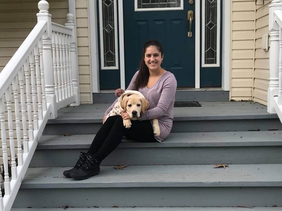 Abby sits on the porch and smiles for the camera with adolescent yellow lab Fergus on her lap.