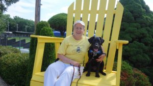 Ruth sits on the oversized yellow outdoor chair located at an outdoor golf driving range. Black lab Wafer wears her blue Guiding Eyes training vest while sitting beside Ruth.
