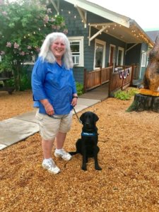 Raiser Kristin Meredith is standing in pea gravel with male black Labrador Dozer in a sit to her left side. He is slightly in front of her left leg. The Cookies & Cream building and a lilac bush are behind them.