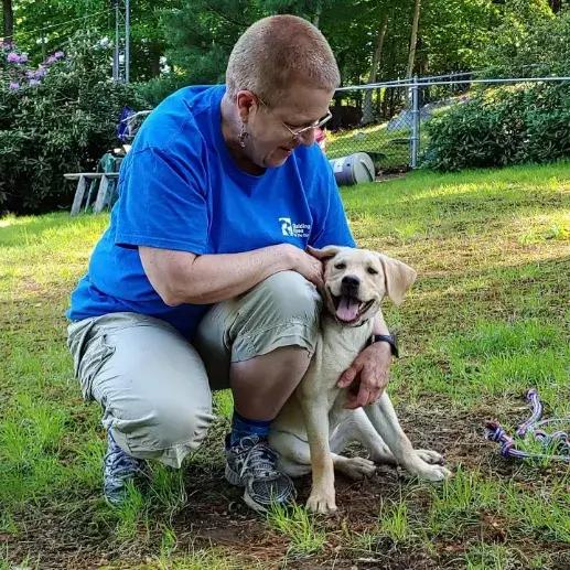 Ruth crouches on the grass beside yellow Labrador retriever puppy Nutmeg. Nutmeg sits beside Ruth and gives a big puppy smile for the camera.