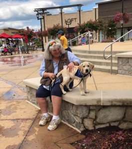 Raiser Kristin Meredith is sitting on the concrete bench in the gathering area of the Promenade at Gainesville with male yellow Labrador, Lux, in a stand position on her left side. She is holding his leash and has her left hand on his side. He is wearing a blue Guiding Eyes for the Blind scarf.
