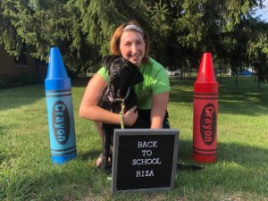 Julia kneels behind adolescent black lab Risa. The pair sit behind a velvet sign board with says "Back To School Risa" and between two crayon decorations, blue and red respectively.