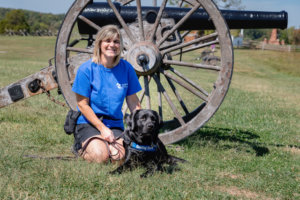 Dawn kneels in the grass with black lab Banner in a down position beside her. Banner wears his blue Guiding Eyes training vest. The pair pose in front of an old war cannon at a museum.