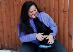 MeKalea smiles down at black lab Alibi. Alibi is wearing her blue Guiding Eyes training vest, and lifts her head to give MeKalea a kiss on the cheek.