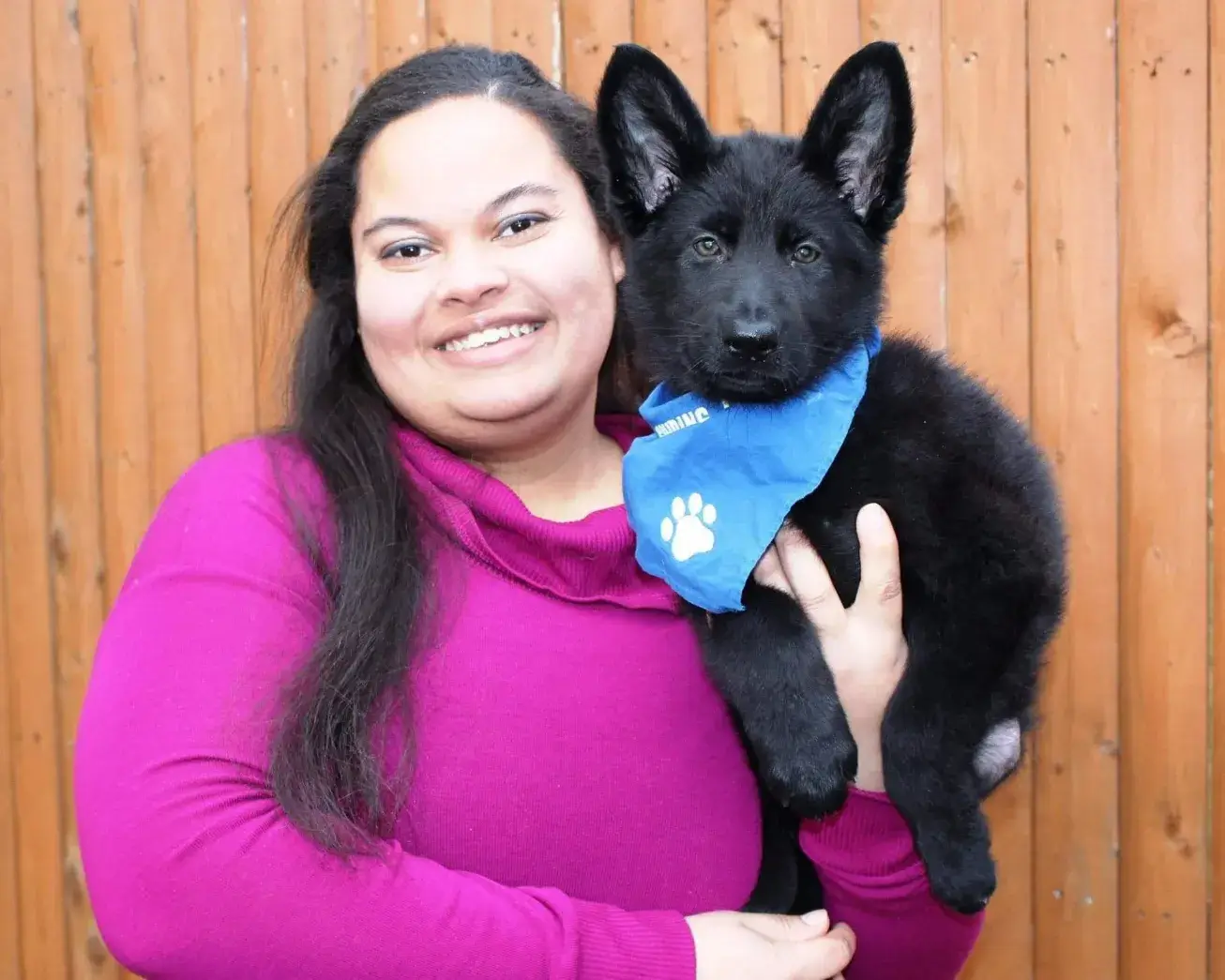 MeKalea holds black German Shepherd puppy Reagan in her arms. Reagan is wearing a blue bandana and looks toward the camera with pricked ears.