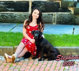 Stephanie Leonor wears a red print dress and sits on a curb with black lab Dover at her side. Stephanie is graduating from Gilmour Academy.