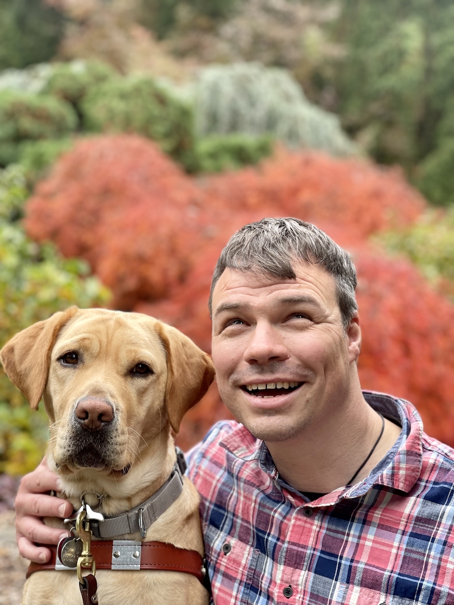 Graduate Adam and guide dog Ursa against a backdrop of orange fall foliage