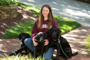 Ainsley sits on the brick path outside surrounded by black labs Tag, Judge, and Kingston.