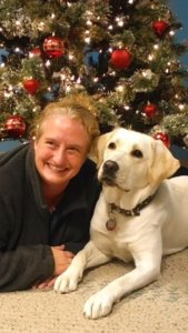 Danielle lays on her stomach on the ground and poses for a photo with yellow lab Annalee in front of a beautifully decorated Christmas tree.