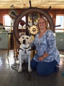 Danielle kneels on the ground beside sitting yellow lab Annalee. The pair pose in front of a large wooden ship helm at a nautical museum outing.