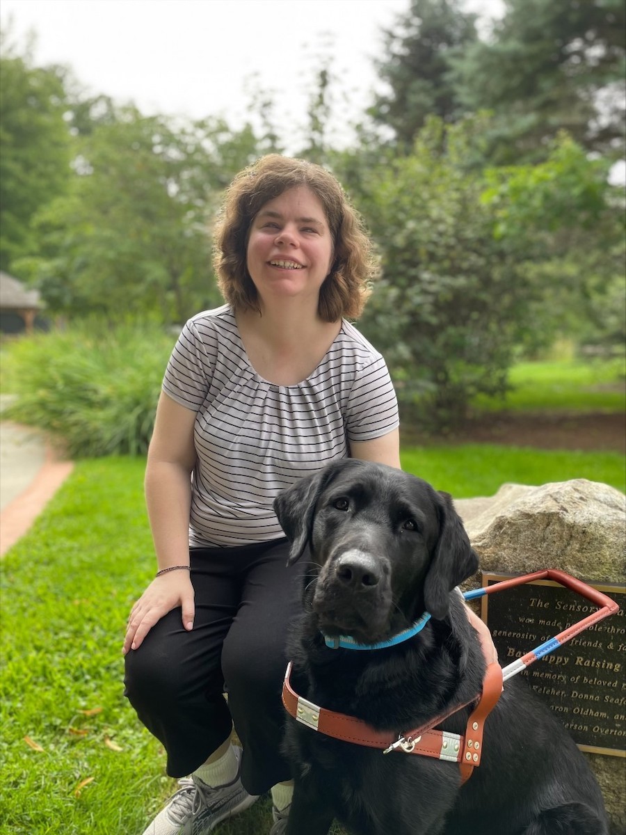 Graduate Ashley poses by a rock with a plaque on campus with guide dog Pendleton