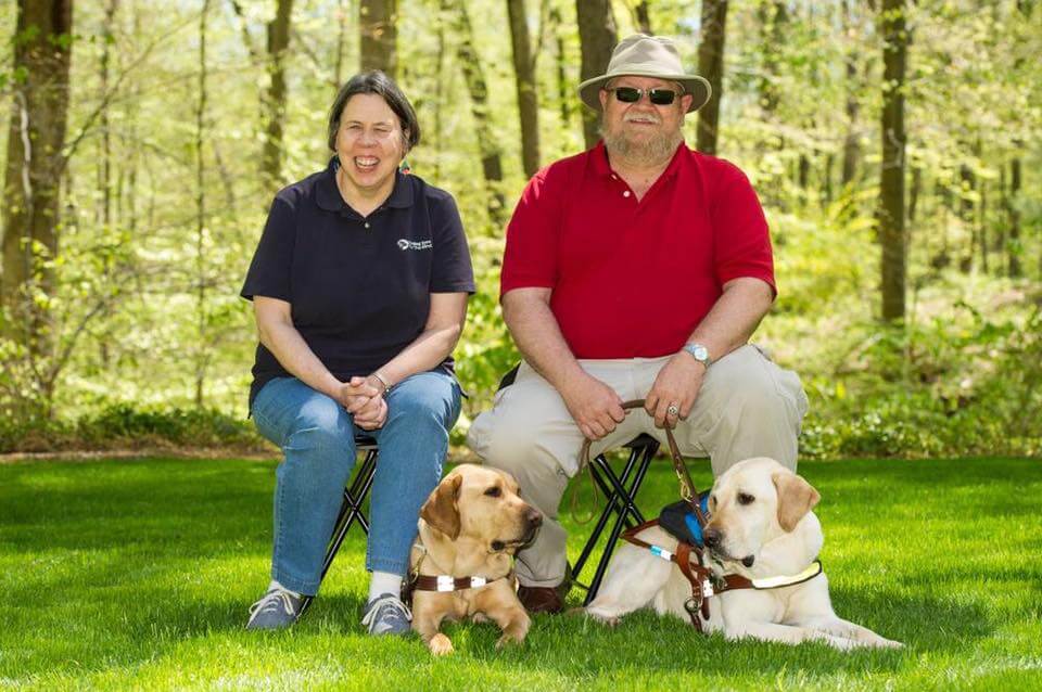 Becky Davidson sits on a bench outside with her husband Ron and guide dogs Lawson and Clarissa