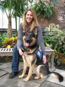 Amy sits on a wooden bench and smiles with pup on program Cappy, a black and tan German Shepherd in a sit at her feet. Cappy wears a blue training vest with the old Guiding Eyes logo.