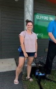 Christina Brilla stands outside a Unity College campus building with black lab Walden laying at her feet. Walden looks up to check in with Christina with his large tongue hanging out of his mouth.