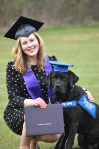 Elizabeth kneels in the grass while wearing her graduation cap and purple stole and holding her graduation diploma out for the camera to see. Black lab Tacoma, wearing a blue graduation cap and her blue pup on program vest, sits next to Elizabeth.