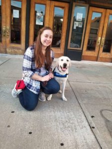 Courtney kneels on the sidewalk beside sitting yellow lab Kipling who is dressed in his blue Guiding Eyes training vest. The pair pose outside of a movie theater before a screening of "Pick of the Litter".