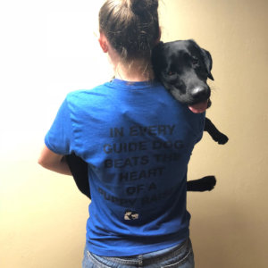 Julia, with her back to the camera, holds black lab Dakota in her arms. Dakota has her head on Julia's shoulder as she looks toward the camera. The back of Julia's blue shirt reads, "In every guide dog beats the heart of a puppy raiser."