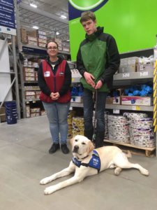 Wes stands in a Lowe's next to an employee with yellow lab Henry laying at Wes' feet. Henry is wearing his blue pup on program vest.