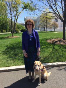 Kathy, dressed up to receive an award for her teaching, stands on the sidewalk outside the White House with yellow lab guide dog Nacho in a sit at her side.