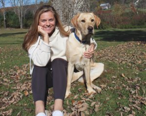 Emily sits in the grass with her hand on her chin and an arm around sitting yellow lab Keene.