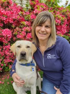 Dawn smiles with her arm around sitting yellow lab Colonel. The pair are in front of some bright pink flowers.