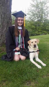 Alexandrea kneels in the grass wearing a graduation cap and gown with yellow lab Vickie at her side.