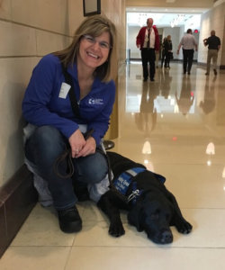 Dawn kneels on the tiled floor in an office building with black lab banner in a down position beside her. Banner wears his blue Guiding Eyes training vest.