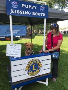 Susan stands next to the Guiding Eyes puppy kissing booth at a lions event. Ipso, a fox red lab, sits in the booth ready for some smooches.
