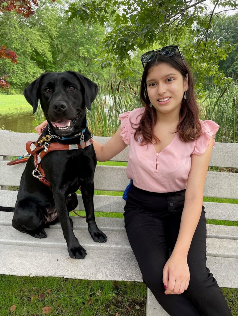 Graduate Joicee and her guide dog Junie, a female black Lab in harness, both sit on the nature path bench at the Guiding Eyes training school campus with the pond in the background. Joicee gently puts her arm around Junie as she smiles toward the camera