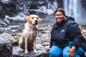 Jessica sits on the rock in front of a waterfall with yellow lab Missy.