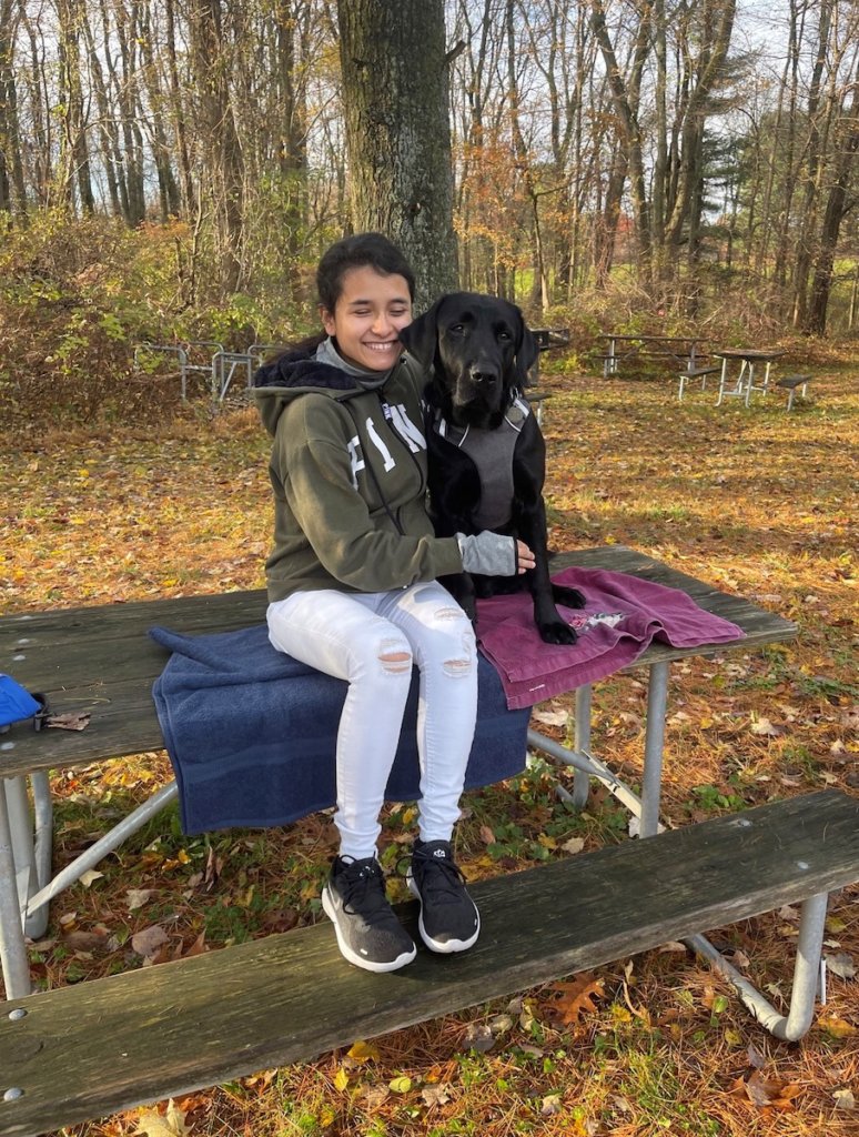Graduate Karlee Karlee and her guide dog Pancho, a male black Lab in harness, both sit on top of a park bench in a fall setting, with an array of different colored leaves covering the ground beneath them. Karlee gently huge Pancho as he glances towards the camera