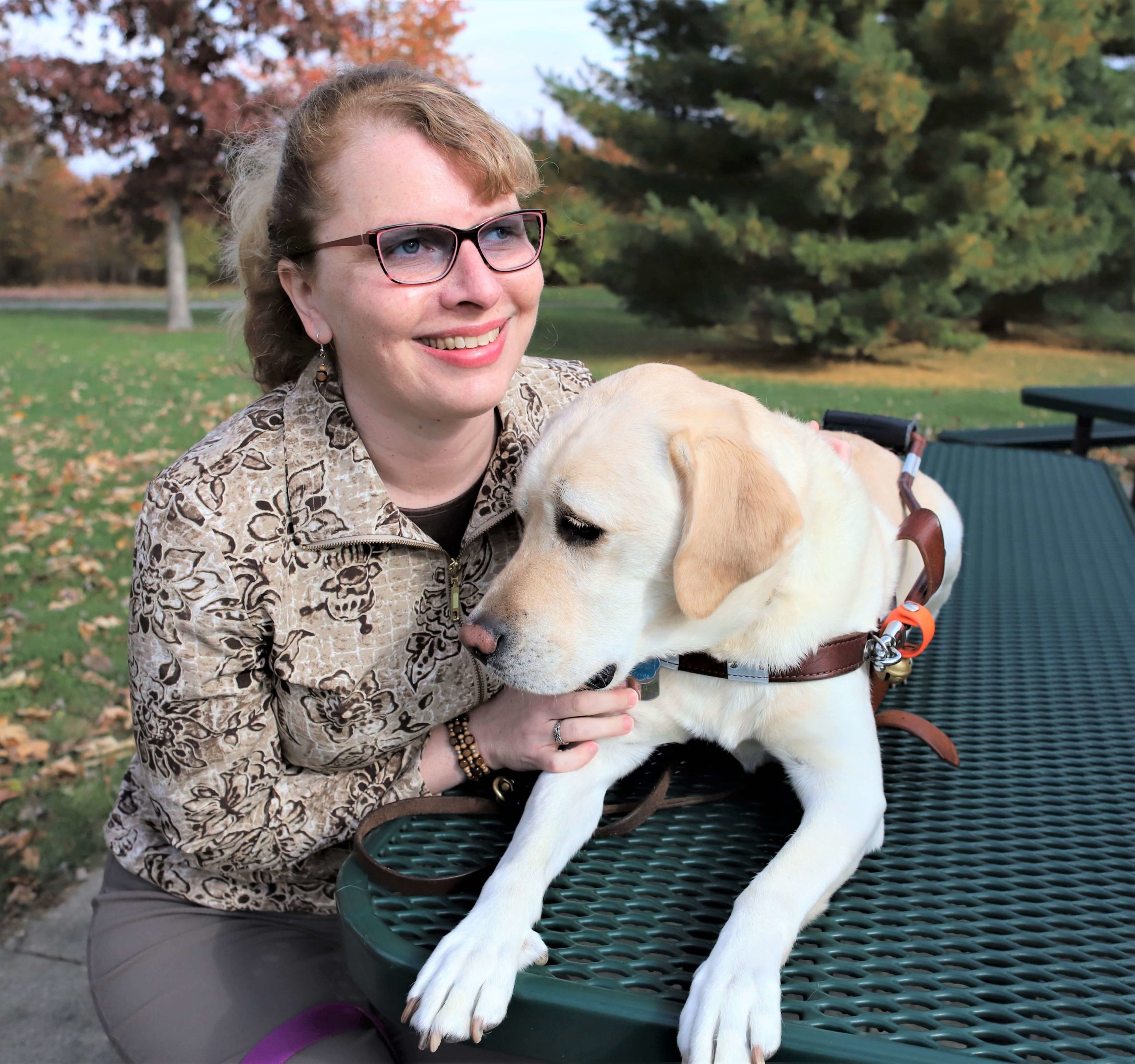 Kathy sits at a park table with yellow lab guide dog Nacho who lays on the grated rubber table.