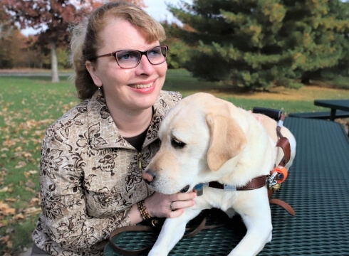 Kathy sits at a park table with yellow lab guide dog Nacho who lays on the grated rubber table.