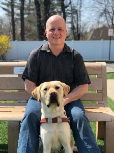 Guide dog Dart sits in front of his handler Kevin on bench on the Yorktown campus