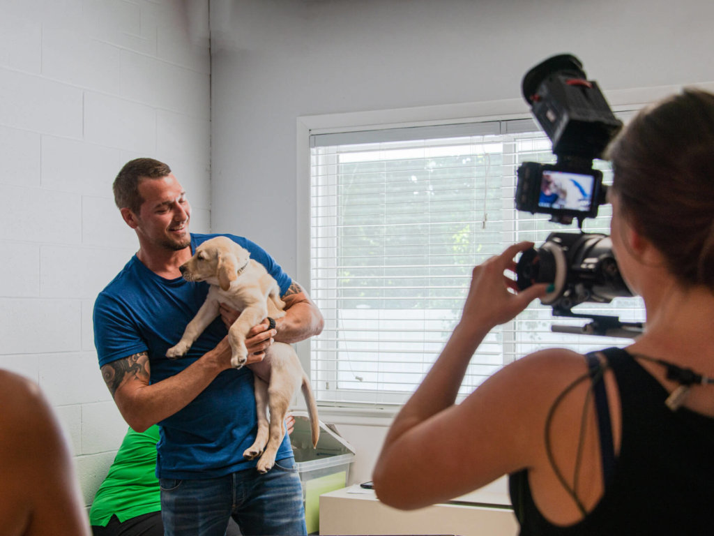 Lucky dog host Brandon snuggles a yellow lab puppy while on camera in the Grooming Room at our Yorktown Heights campus.