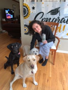Hannah Schayes sits on a chair with a graduation banner hung on the wall in the background. Hannah holds a 2020 graduation coffee mug in her hand as she smiles and bends over to pose with a photo with her pack of dogs: black lab Junie (on program), yellow lab Vera (released), and scruffy white pup Oliver (family pet).