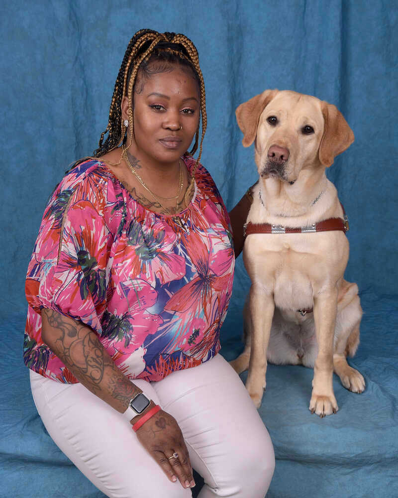 Navonne and her guide dog Praline, a female yellow Lab in harness, both sit in front of a denim blue background. Navonne lays her hand gently around Praline as she stares towards the camera