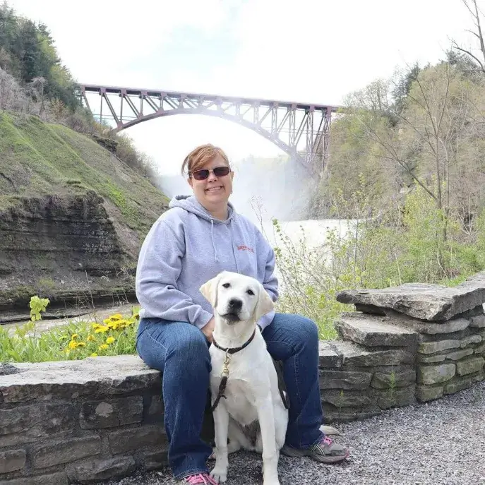 Renee sits on the low stone wall at a National Park with a bridge over a river in the distance. At Renee's feet sits yellow lab Fortune.