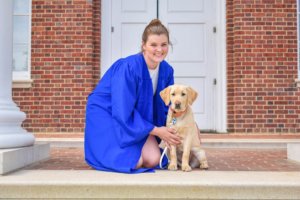 Olivia Miller kneels on the ground outside a campus building wearing a blue graduation gown. Young yellow lab pup Cici sits beside her and looks eagerly toward the camera.