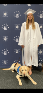 Paige stands in her white graduation cap and gown with yellow lab Bo in a down position at her feet. The pair stand in front of a blue step-and-repeat patterned with the logos of Hudson High School.
