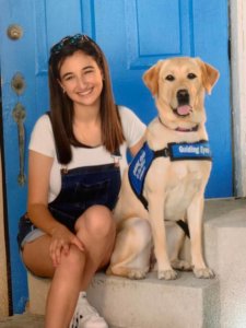 Bryanna Schleifer sits in front of a blue door with yellow Lab Windy. Windy wears her Guiding Eyes puppy jacket. Bryanna attended Orange High School.