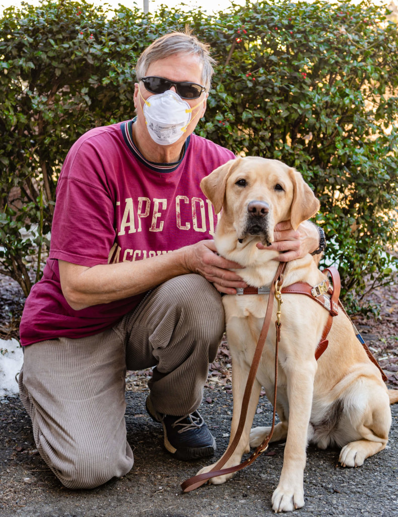 Behind the mask, graduate Phil kneels next to guide dog Kit on a warm spring day of training