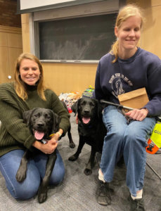 Sam kneels on the carpeted floor of the classroom at Ithaca College with black lab Elaine on her lab. Seat in a chair is Liz Bottner with her guide dog Ocala, a black lab, seated beside her.